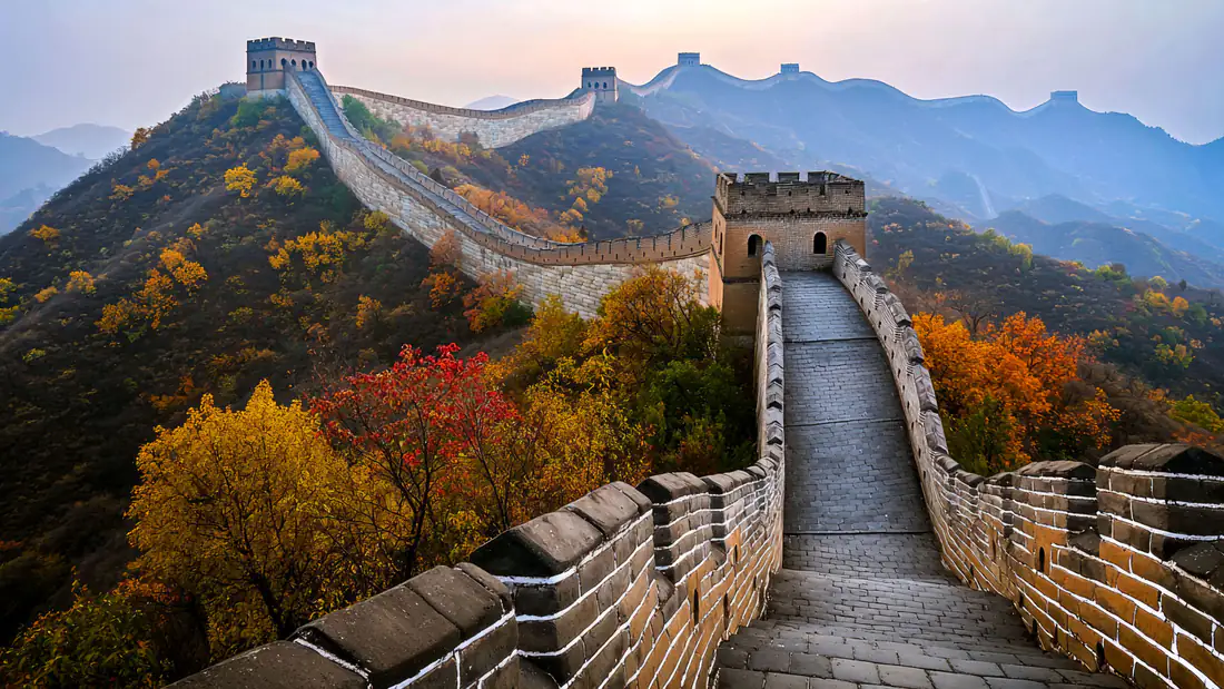 The Great Wall of China winding across forested hills in autumn colours under a hazy sunrise