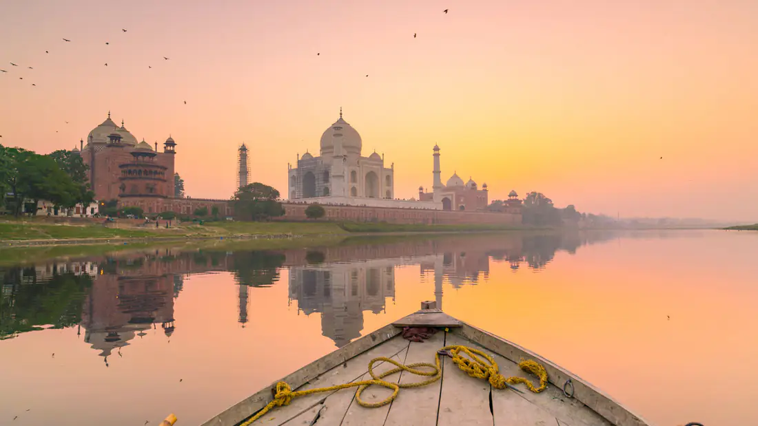 The Taj Mahal at sunrise reflected in the calm Yamuna river, seen from the bow of a wooden boat in Agra, India