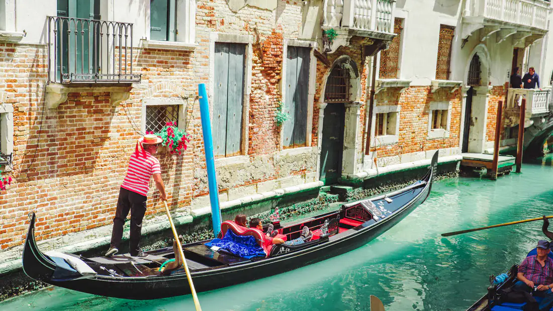 A gondolier in a striped shirt steering a gondola with passengers along a narrow Venice canal past brick buildings