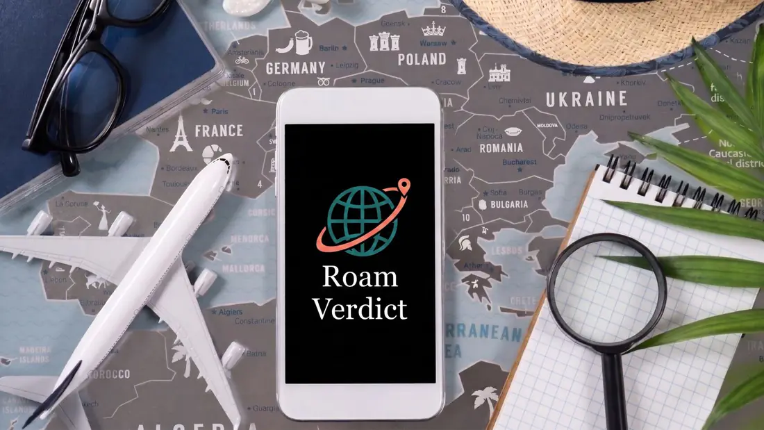 A phone showing the RoamVerdict logo on a travel flat lay with a map of Europe, a model plane, sunglasses, a straw hat and a notebook