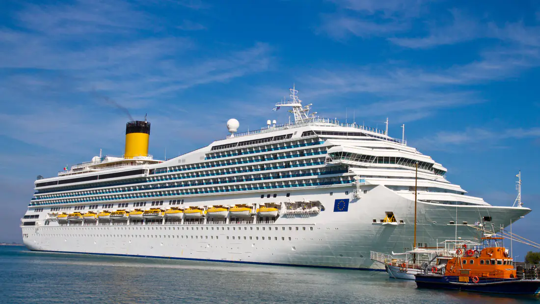 A large white cruise ship moored at port beside a small orange pilot boat under a blue sky