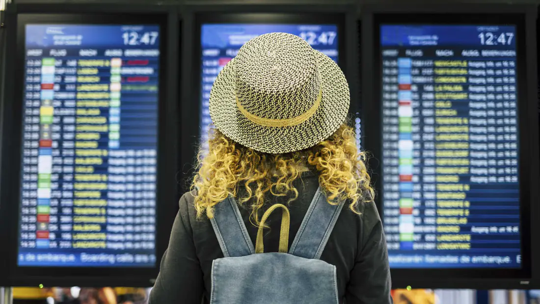 A traveler in a straw hat with a backpack reading European airport departure screens listing flights to Lisbon, Munich, Madrid and Brussels