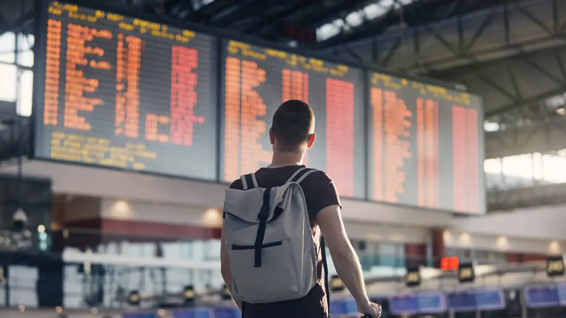 A traveler with a backpack and suitcase looking up at a large amber airport departure board in a terminal